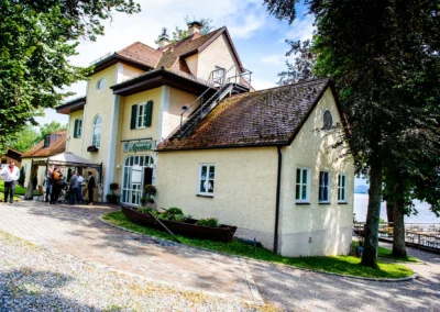 Hochzeitsfeier im Restaurant Alpenblick am Staffelsee mit Gaesten vor dem Gebaeude bei Sonnenschein.