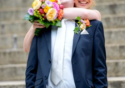 Brautpaar umarmt sich lachend mit Blumenstrauss auf Treppe. Hochzeitsfoto in Muenchen Schwabing.