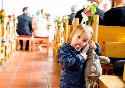 Kleines Kind mit Schnuller und Kuscheltier in einer Kirche bei der Hochzeit in Feldafing, Brautpaar unscharf im Hintergrund.