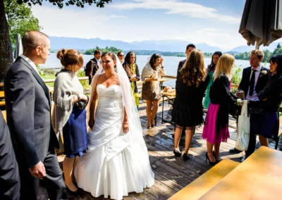 Braut und Gaeste bei Hochzeit am Staffelsee, festliche Atmosphaere mit Blick aufs Wasser und Berge im Hintergrund.