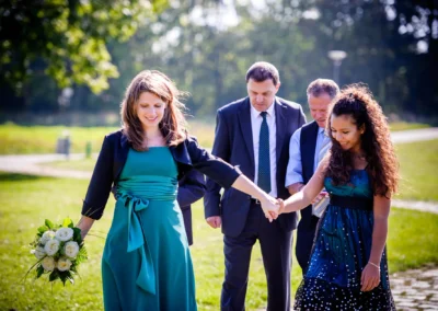 Hochzeitsfoto in Obermenzing: Zwei Frauen in eleganten Kleidern laufen über eine Wiese, begleitet von zwei Männern in Anzügen.