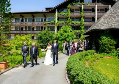 Brautpaar und Hochzeitsgesellschaft spazieren bei sonnigem Wetter durch eine Gartenanlage nahe Grainau.