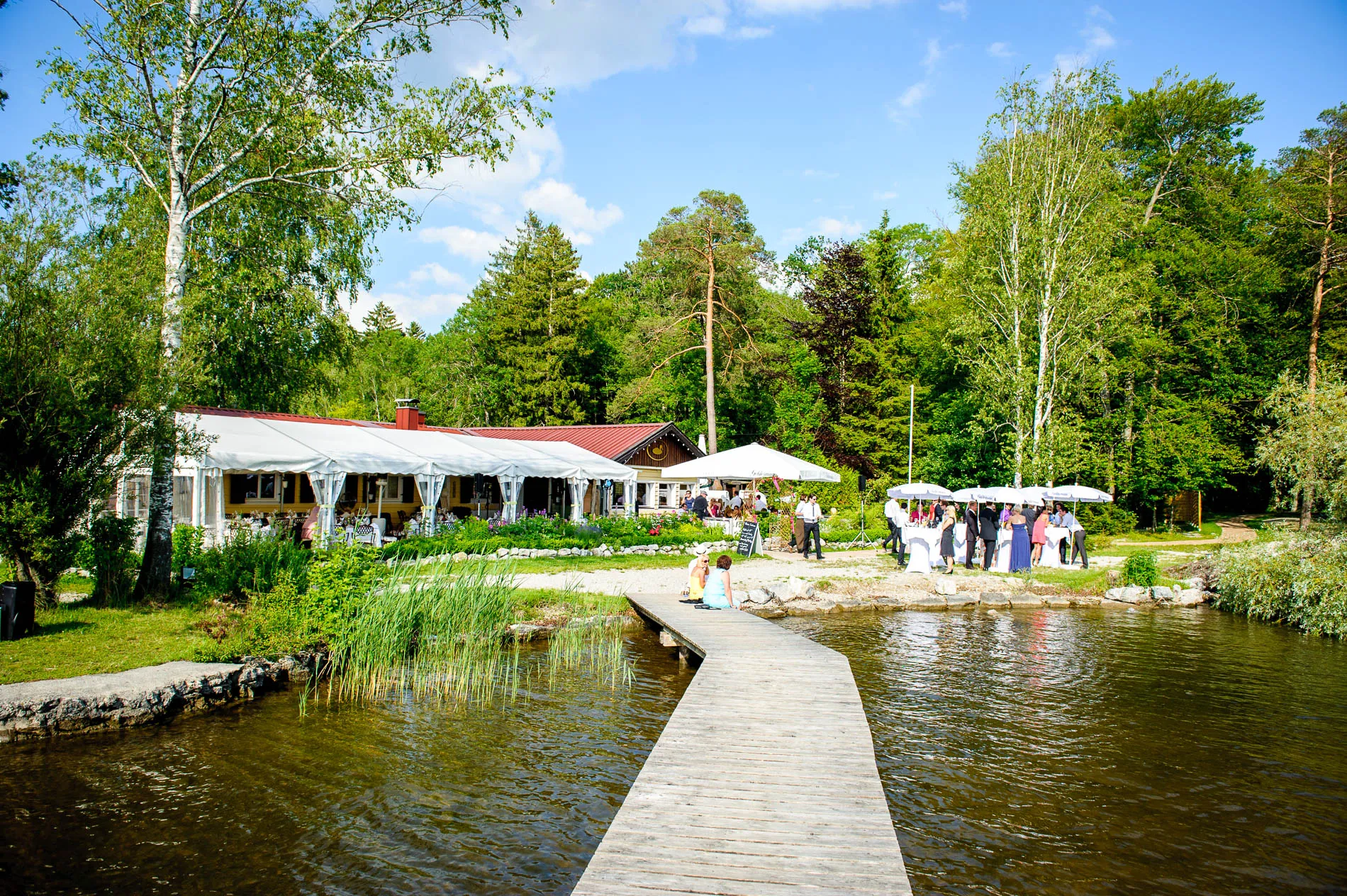 Hochzeitsreportage am Starnbergersee: Seesteg, Feier im Garten unter Pavillons mit Baeumen und See im Hintergrund.