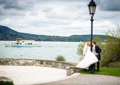 Brautpaar am Tegernsee posiert neben Laterne, im Hintergrund ein Fahrgastschiff auf dem See. Hochzeitsfotografie.