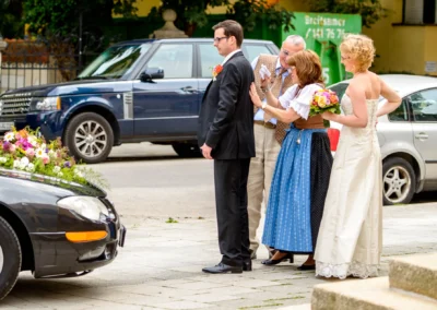 Hochzeitsfoto: Brautpaar mit Familie in Tracht vor Auto mit Blumenschmuck, aufgenommen in München Schwabing.