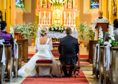 Brautpaar waehrend kirchlicher Trauung in Muenchen. Hochzeitsfotografie mit Blumenschmuck und Priester am Altar.