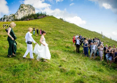Braut begleitet von Gästen auf grüner Bergwiese bei Kampenwand Hochzeit, Fotograf für Hochzeitsreportage.