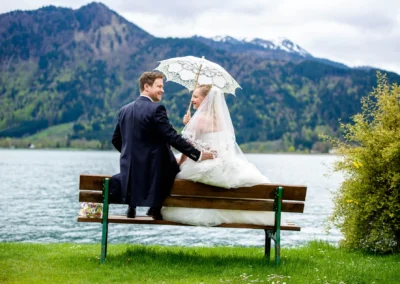 Brautpaar sitzt auf einer Bank am Tegernsee, Braut mit Schirm, im Hintergrund Berge. Hochzeitsfoto Tegernsee.