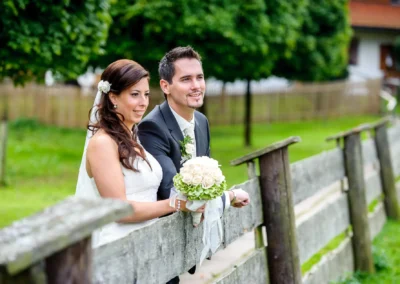 Brautpaar bei Hochzeitsfotos in Sauerlach-Arget, lehnt am Holzzaun mit Blumenstrauss, Hochzeitsszene im Freien.