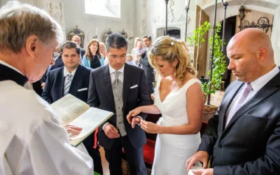 Brautpaar beim Ringtausch in einer Kirche, Priester liest aus Buch, mehrere Gäste im Hintergrund, helle Lichtstimmung.