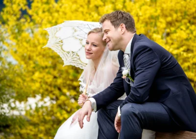 Brautpaar beim Fotoshooting mit Spitzen-Regenschirm vor gelber Blumenpracht am Tegernsee, Hochzeitsfotografie.