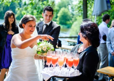 Braut mit Blumenstrauss nimmt Sekt am Staffelsee. Hochzeitsfotografie in Uffing. Fröhliche Aufnahme bei einer Hochzeit.