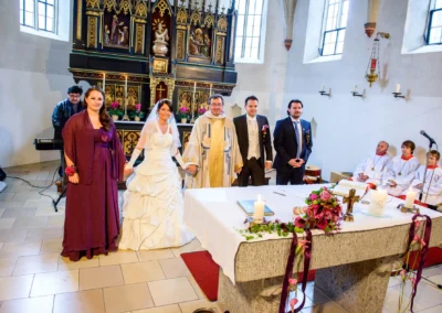 Brautpaar und Priester bei kirchlicher Trauung in St. Vitus Gilching, umgeben von Familie und Freunden.
