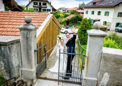 Brautpaar lacht und winkt bei Hochzeit in Waakirchen, umgeben von malerischen Hausern und gruenem Garten.