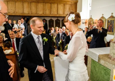 Brautpaar tauscht Ringe waehrend kirchlicher Trauung bei Hochzeit in Gut Thurnsberg, Gaeste beobachten im Hintergrund.