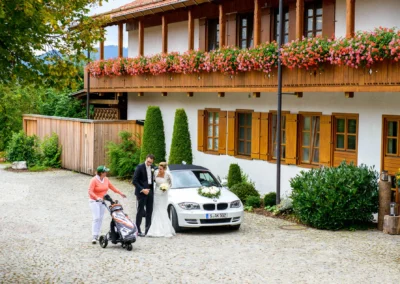 Brautpaar vor Blumenhaus in Waakirchen, aufgenommen bei Hochzeit. WeiBes Auto und Golftasche im Hintergrund. Hochzeitsfoto.