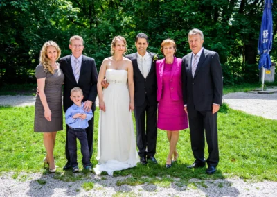 Familienfoto bei Hochzeit im Luitpoldpark Muenchen. Brautpaar in der Mitte, umgeben von Gästen bei Sonnenschein.