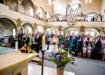 Kirchliche Trauung in der Erloeserkirche Schwabing, Brautpaar und Gaeste stehen vor dem Altar mit Blumenarrangement.