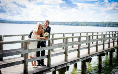 Brautpaar auf Holzsteg über Wasser, sie hält Blumenstrauß, er umarmt sie, sonniges Wetter, bewölkter Himmel im Hintergrund.