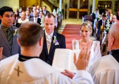 Brautpaar bei kirchlicher Trauung in Muenchen, Hochzeitsfotografie in der Kirche mit Priester und Gaesten im Hintergrund.