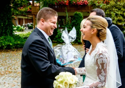 Brautpaar lacht und tauscht Geschenk aus bei Hochzeitsreportage nahe Tegernsee. Hochzeit mit Blumen im Hintergrund.
