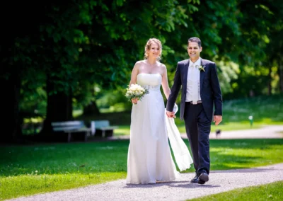 Brautpaar spaziert im Luitpoldpark Muenchen, sie traegt ein weisses Kleid und haelt Blumenstrauss. Hochzeitsreportage.