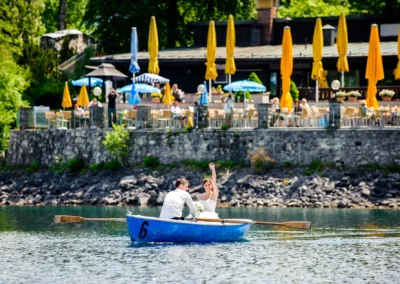 Brautpaar in blauem Boot auf See nahe Grainau bei Hochzeit, im Hintergrund Restaurant mit gelben Sonnenschirmen.