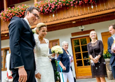 Brautpaar lacht vor einem blumengeschmückten Haus in Waakirchen bei ihrer Hochzeit. Hochzeitsreportage in Bayern.