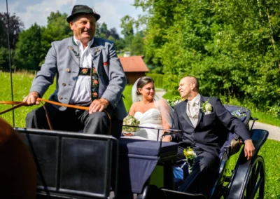 Brautpaar in Kutsche mit Kutscher bei Hochzeitsreportage in Uffing am Staffelsee, umgeben von gruenen Baeumen.