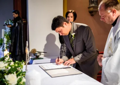 Ein Mann unterschreibt waehrend der kirchlichen Trauung in Muenchen, begleitet von einer Frau und einem Priester.