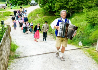 Hochzeit in Oberbayern: Ein Mann mit Akkordeon fuehrt Gaeste in traditioneller Tracht an. Hochzeitsfotografie.