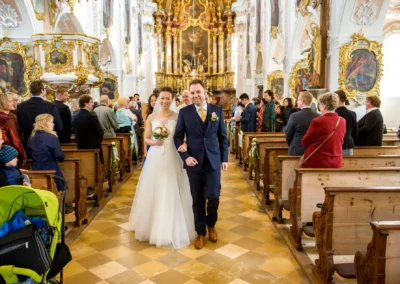 Brautpaar laechelt nach kirchlicher Trauung in Kirche nahe Fischbachau, Hochzeitsfotografie mit Gaesten und prunkvollem Altar.