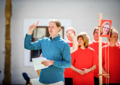 Hochzeitsreportage nahe des Chiemsees: Mann in blauem Hemd hebt Hand, Personen in Rot lachen im Hintergrund.