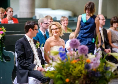 Brautpaar und Gaeste bei kirchlicher Trauung in Schwabing. Blumenarrangement im Vordergrund. Hochzeitsfotografie.