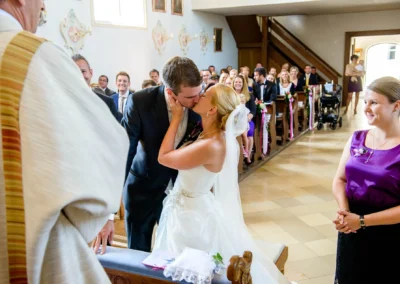 Brautpaar bei kirchlicher Hochzeit in Taufkirchen; Hochzeitsfoto mit Kuss vor Altar und Gästen.