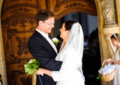 Brautpaar laechelt sich vor Holztuer an, Maedchen mit Korb daneben. Kirchliche Trauung in Aufkirchen am Starnberger See.