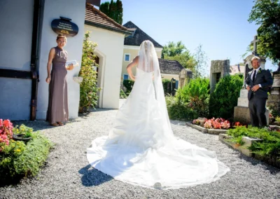 Braut in weissem Kleid vor Kirche in Bernried, begleitet von Gästen bei sonnigem Wetter. Hochzeitsfotografie.