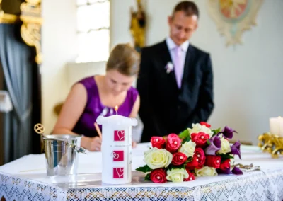 Kerze und Blumenstrauß auf Tisch bei kirchlicher Trauung in Kirche nahe München, Paar im Hintergrund. Hochzeitsfoto.