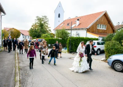 Brautpaar bei kirchlicher Trauung in Gilching, gefolgt von Gästen, die durch die Straße zur St. Vitus Kirche spazieren.