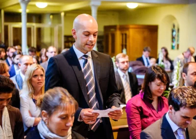 Mann liest in Kirche waehrend einer Hochzeit in Muenchen, Gaeste aufmerksam. Hochzeitsfotografie in Pasing.