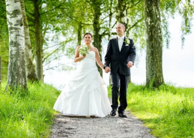 Brautpaar auf Waldweg bei Hochzeit am Staffelsee, umgeben von hohen Baeumen, Hochzeitsreportage mit natuerlichem Licht.