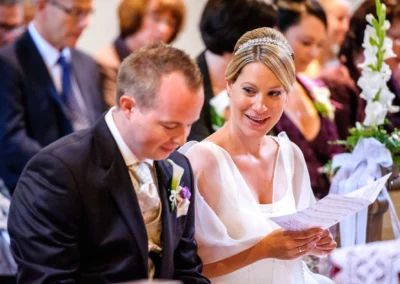 Brautpaar bei kirchlicher Trauung in Muenchen, sie laechelt mit Programmheft, umgeben von Gaesten. Hochzeitsfotograf.