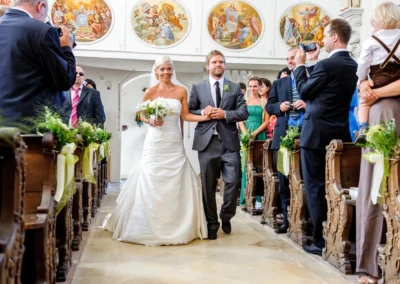 Braut schreitet mit Begleiter durch Kirchgang in Starnberg, Brautstrauss in Hand. Hochzeitsfotografie kirchliche Trauung.