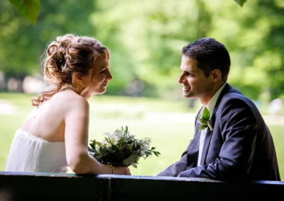 Brautpaar bei Hochzeit im Luitpoldpark Muenchen, Braut mit Blumenstrauss und laechelnder Bräutigam in eleganten Anzug.
