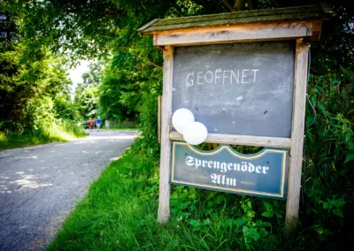 Hinweisschild mit Geoeffnet bei der Sprengenoeder Alm, umgeben von Gruen, ideal fuer Hochzeitsfotografie.