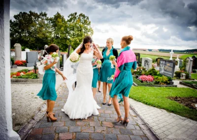 Braut mit Brautjungfern bei kirchlicher Trauung in Arget. Hochzeitsfotografie im Freien zwischen Gruenanlagen und Grabsteinen.
