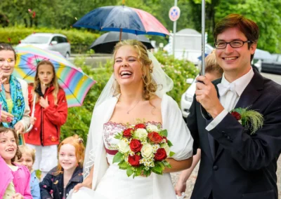 Brautpaar lachend mit Blumenstrauss bei einer Hochzeit am Walchensee, umgeben von Gästen mit Regenschirmen.