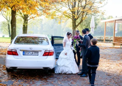 Braut im weissen Kleid und Blumenstrauss vor Auto bei einer Hochzeitsreportage im Paulaner Seegarten.