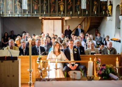 Kirchliche Trauung in Muenchen, Brautpaar sitzt in Kirche umgeben von Gaesten. Hochzeitsreportagefotografie.
