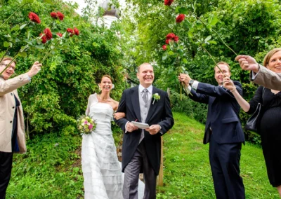 Brautpaar laechelt in die Kamera, umgeben von Gaesten mit Rosen, bei einer Hochzeitsreportage auf Gut Thurnsberg.